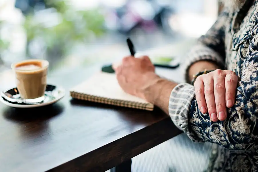 Close up image of an elderly author's hand writing in a notebook. The author is sitting at a table with an iced coffee next to their work.
